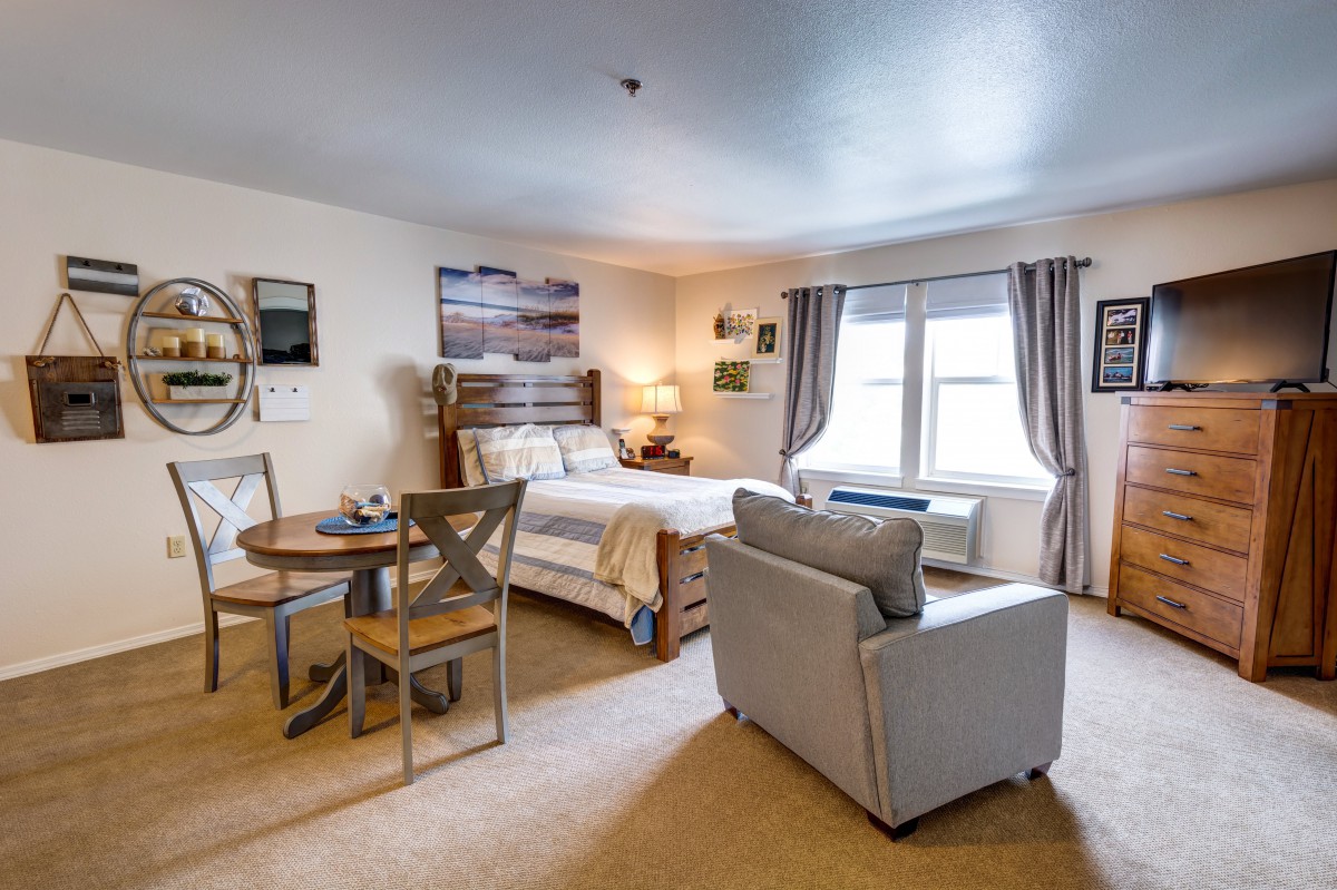 A cozy studio apartment features a comfortable bed, a small dining area, and ample natural light.