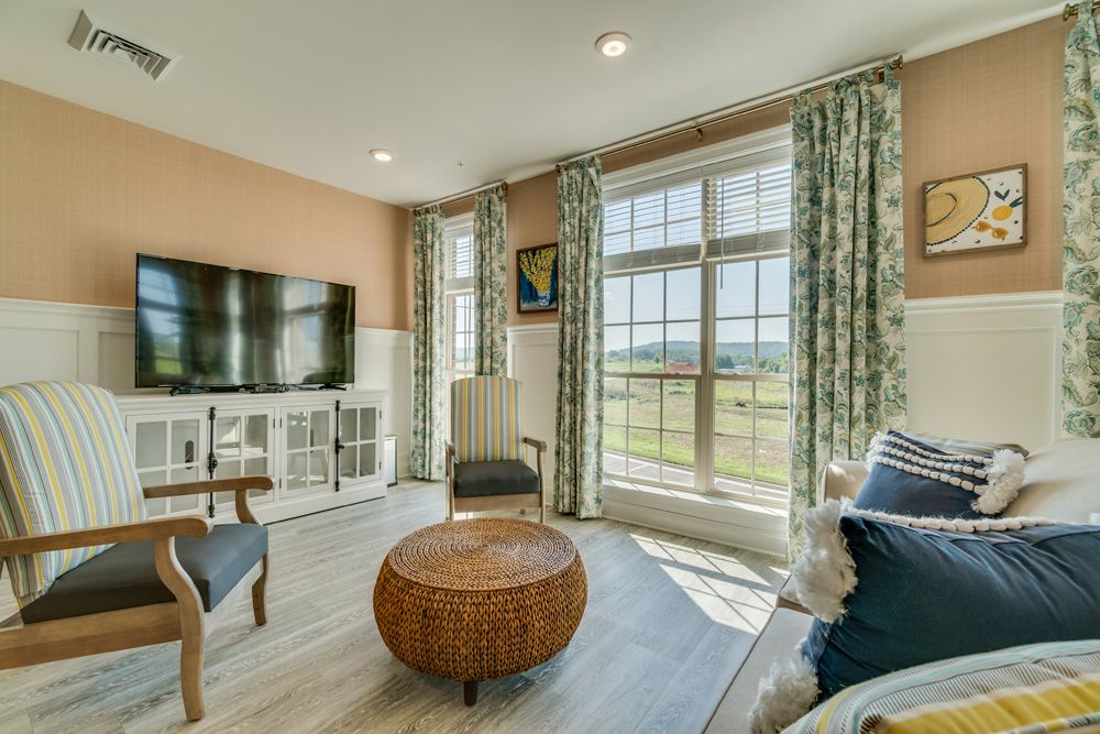 A bright living room featuring large windows with floral curtains, a mounted television, and comfortable seating.