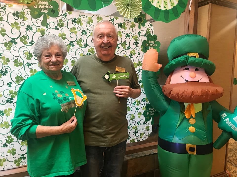 An elderly couple poses with a leprechaun cutout during a St. Patrick's Day celebration, surrounded by festive decorations.