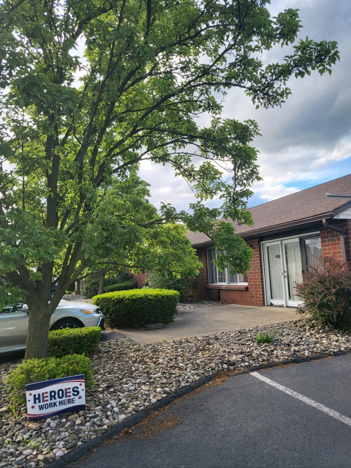 A suburban house with a tree in the yard and a driveway leading to parked cars under a cloudy sky.