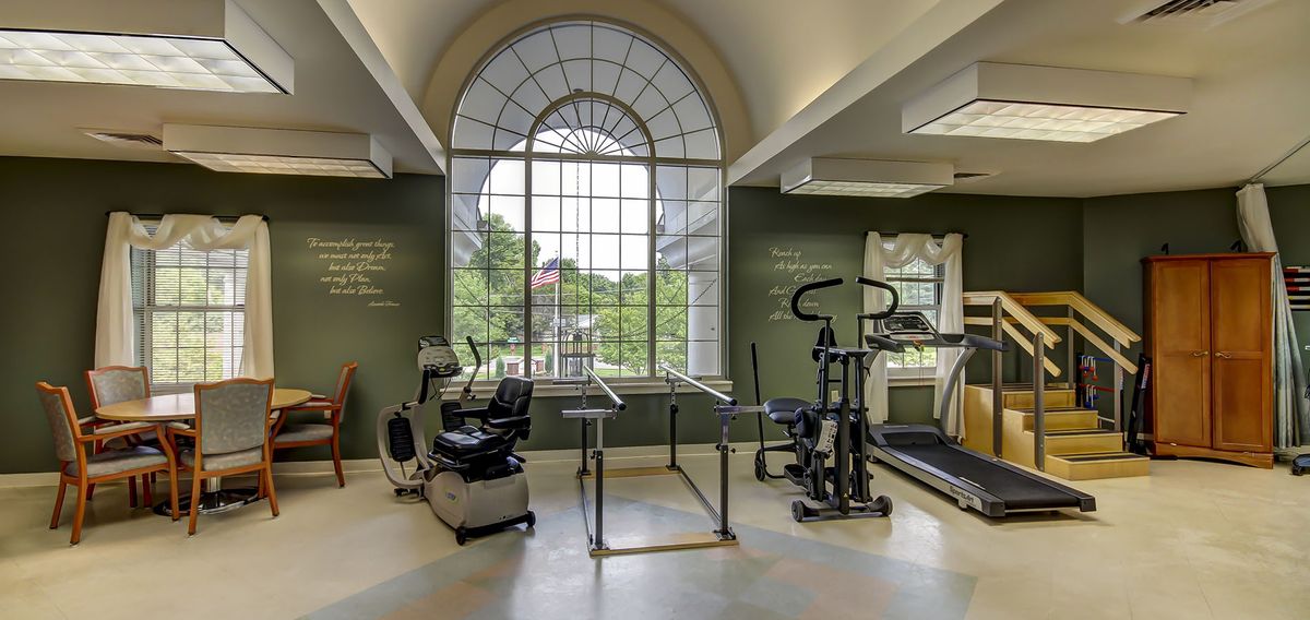A well-lit gym with various exercise equipment in front of a large arched window.