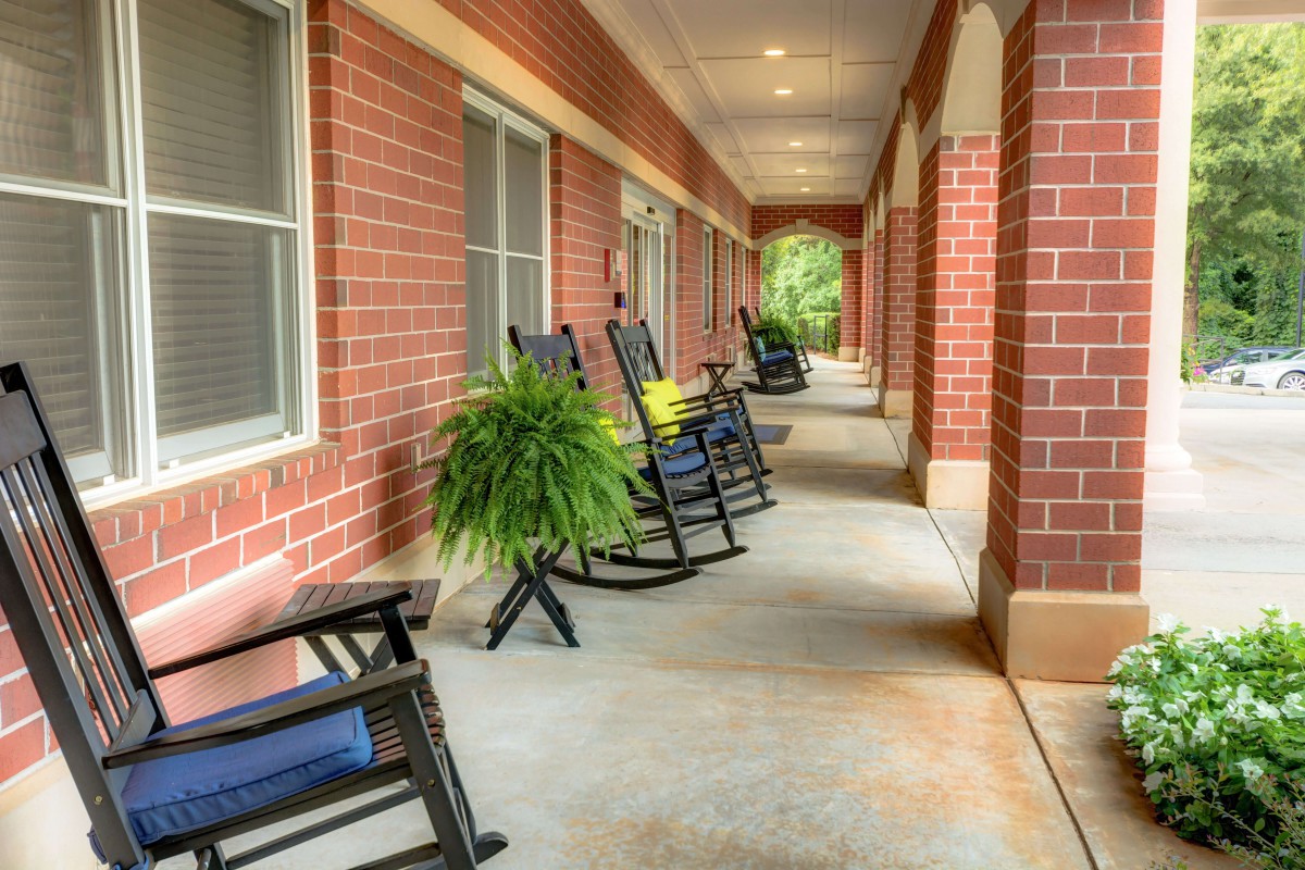 A serene porch with rocking chairs and lush plants along a brick building corridor.