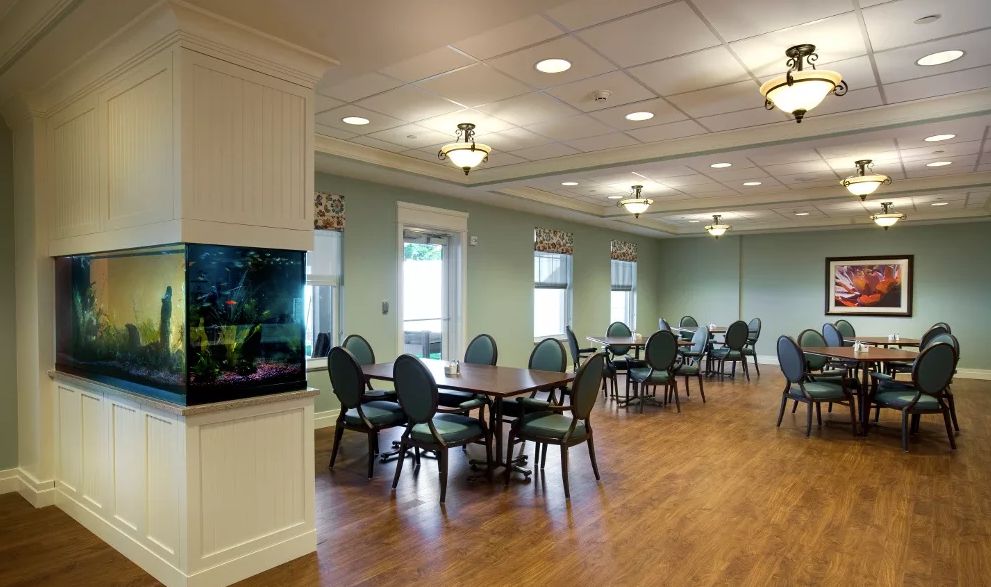 A well-lit dining area with multiple tables and chairs, featuring a television on the wall and wooden flooring.