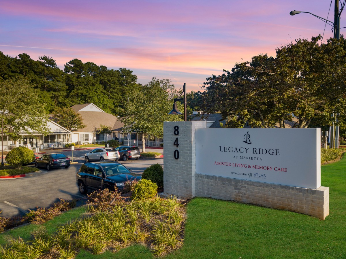 A view of Legacy Ridge, a retirement community, with its sign in the foreground and building structures surrounded by trees at dusk.