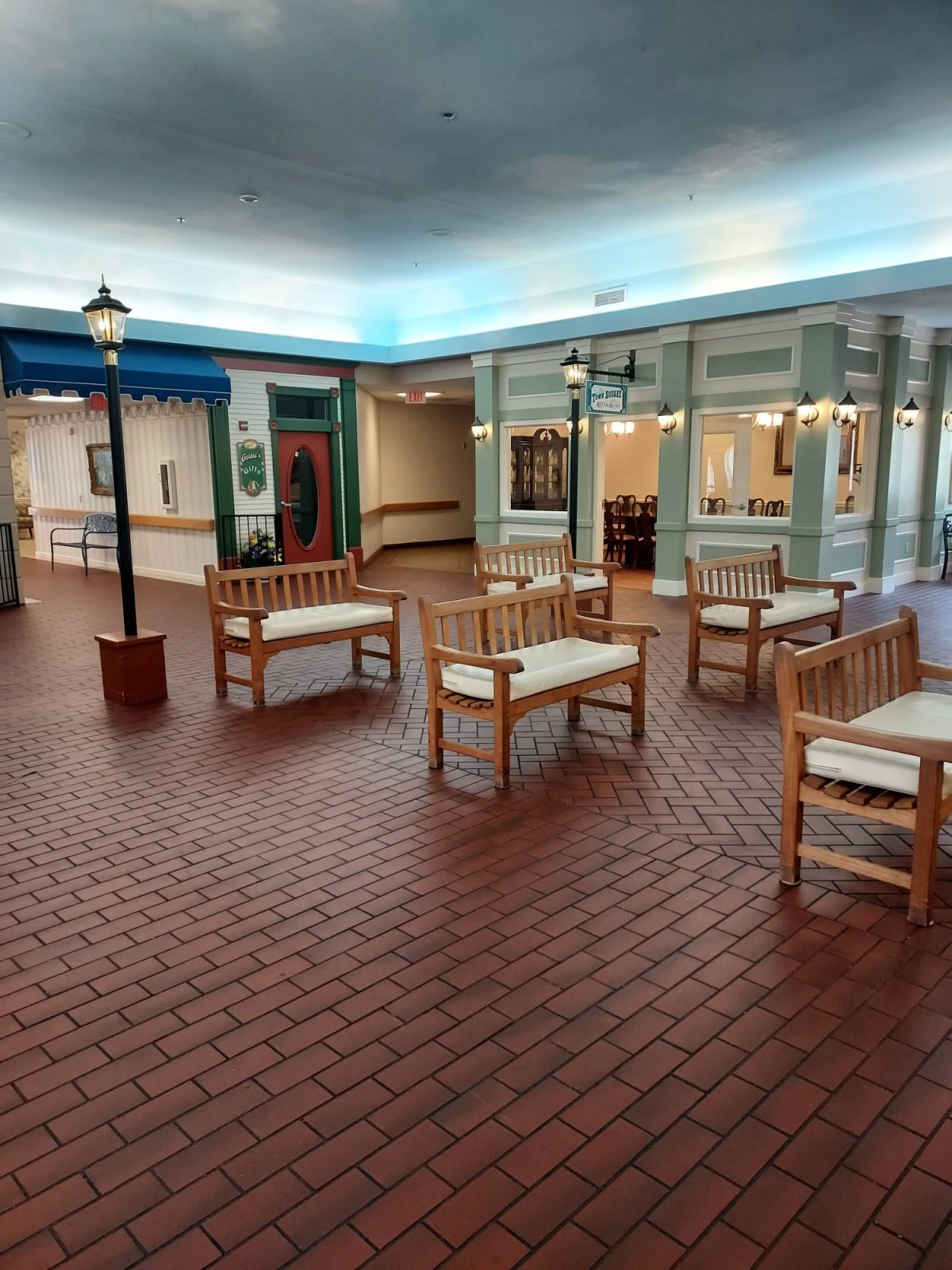 An indoor seating area with benches and brick flooring, featuring street lamps and a storefront-like facade.