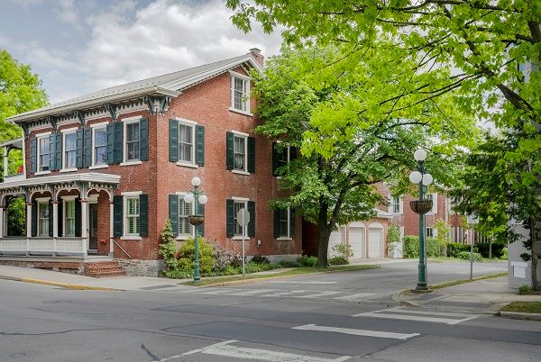 A charming brick house on a quiet street corner surrounded by lush green trees.