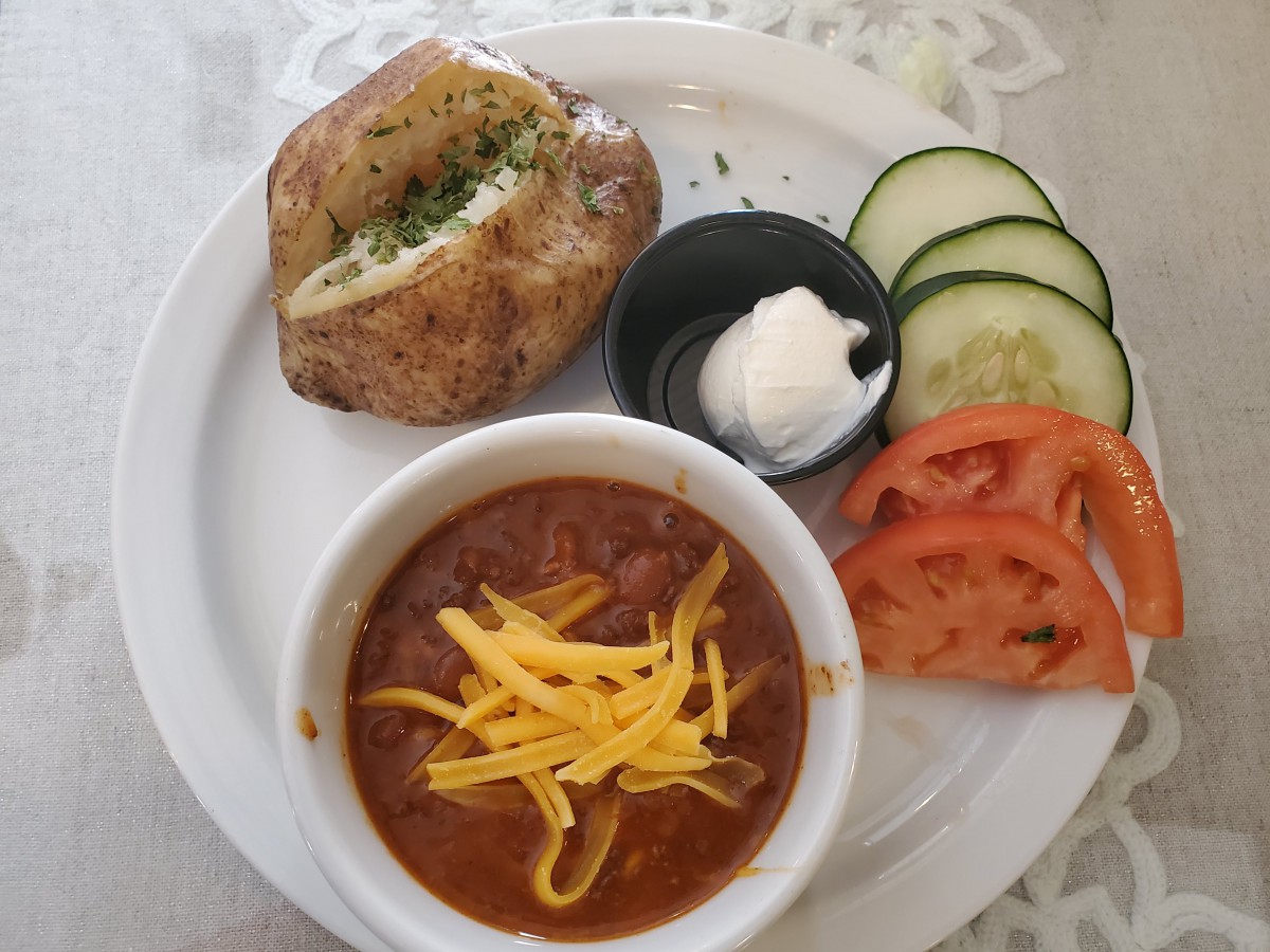 A plate featuring a bowl of chili topped with cheese, accompanied by a baked potato, cucumber and tomato slices, and a side of sour cream.