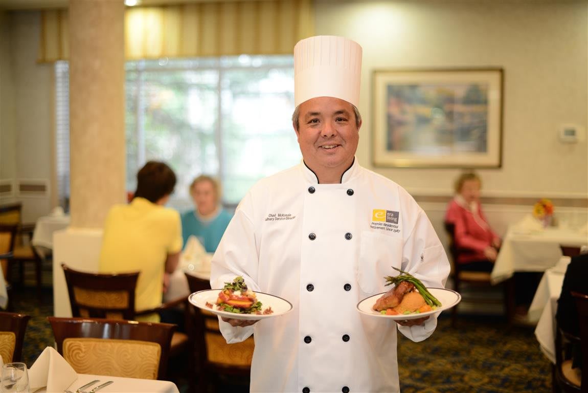 A chef in a professional kitchen holding two plates of food with a smile.