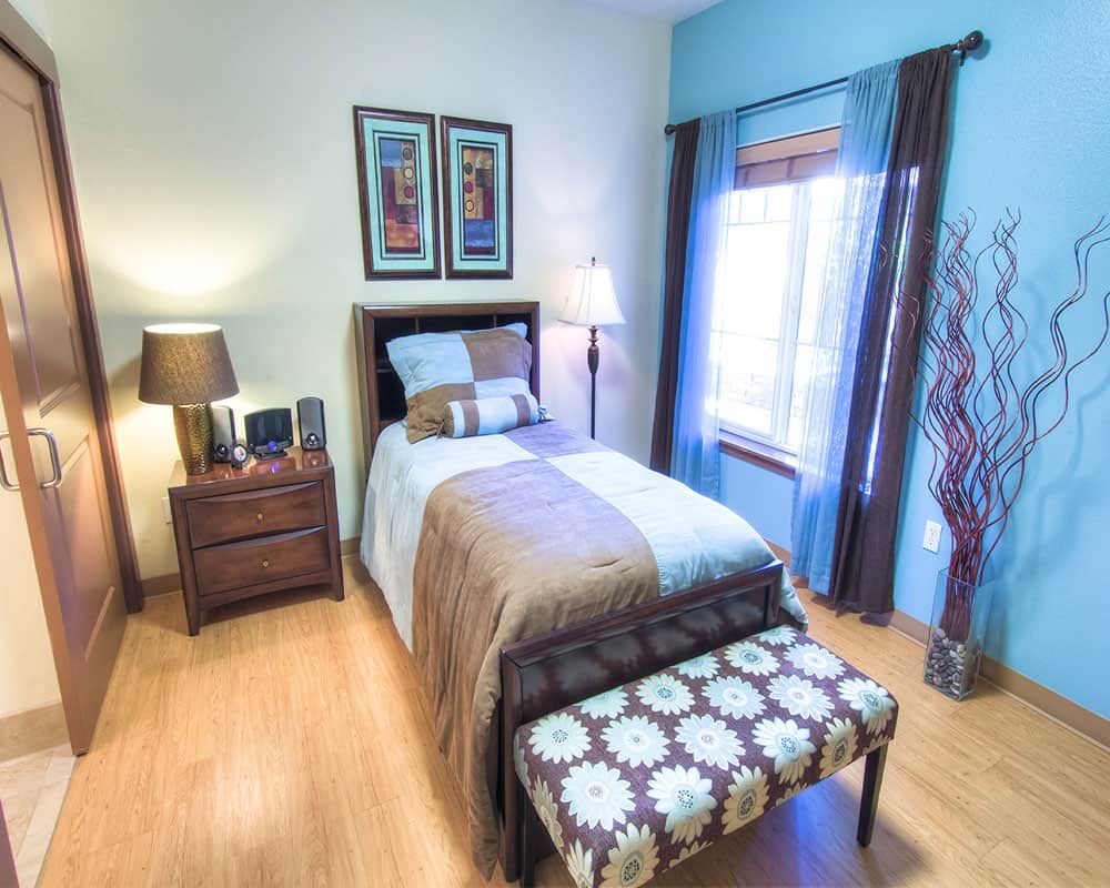 A cozy bedroom with modern decor, featuring a single bed, side table, lamp, and large window.