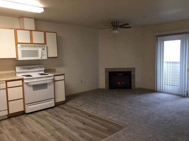 A cozy apartment living space featuring a kitchen with white cabinets and appliances, alongside a fireplace and ceiling fan.