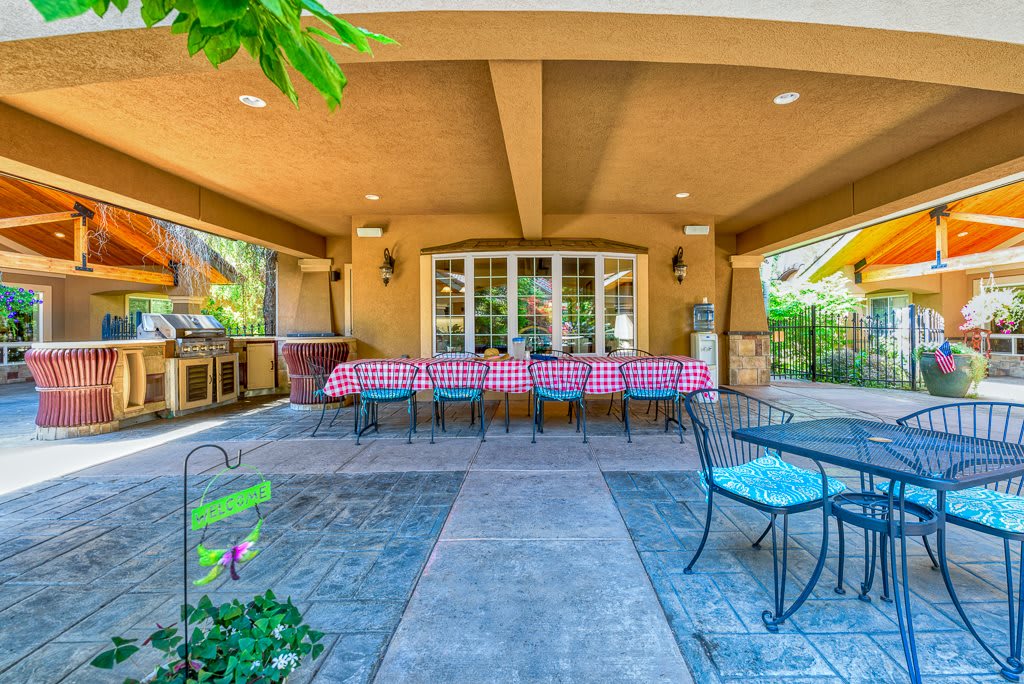 A spacious outdoor patio with a long dining table and checkered tablecloth, surrounded by chairs under a covered roof.