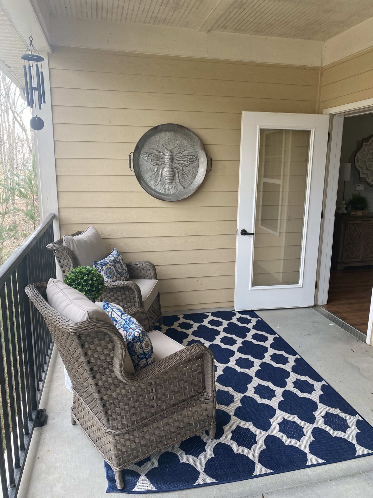 A cozy balcony with wicker chairs, decorative pillows, a potted plant, and a patterned rug leading to an open door.