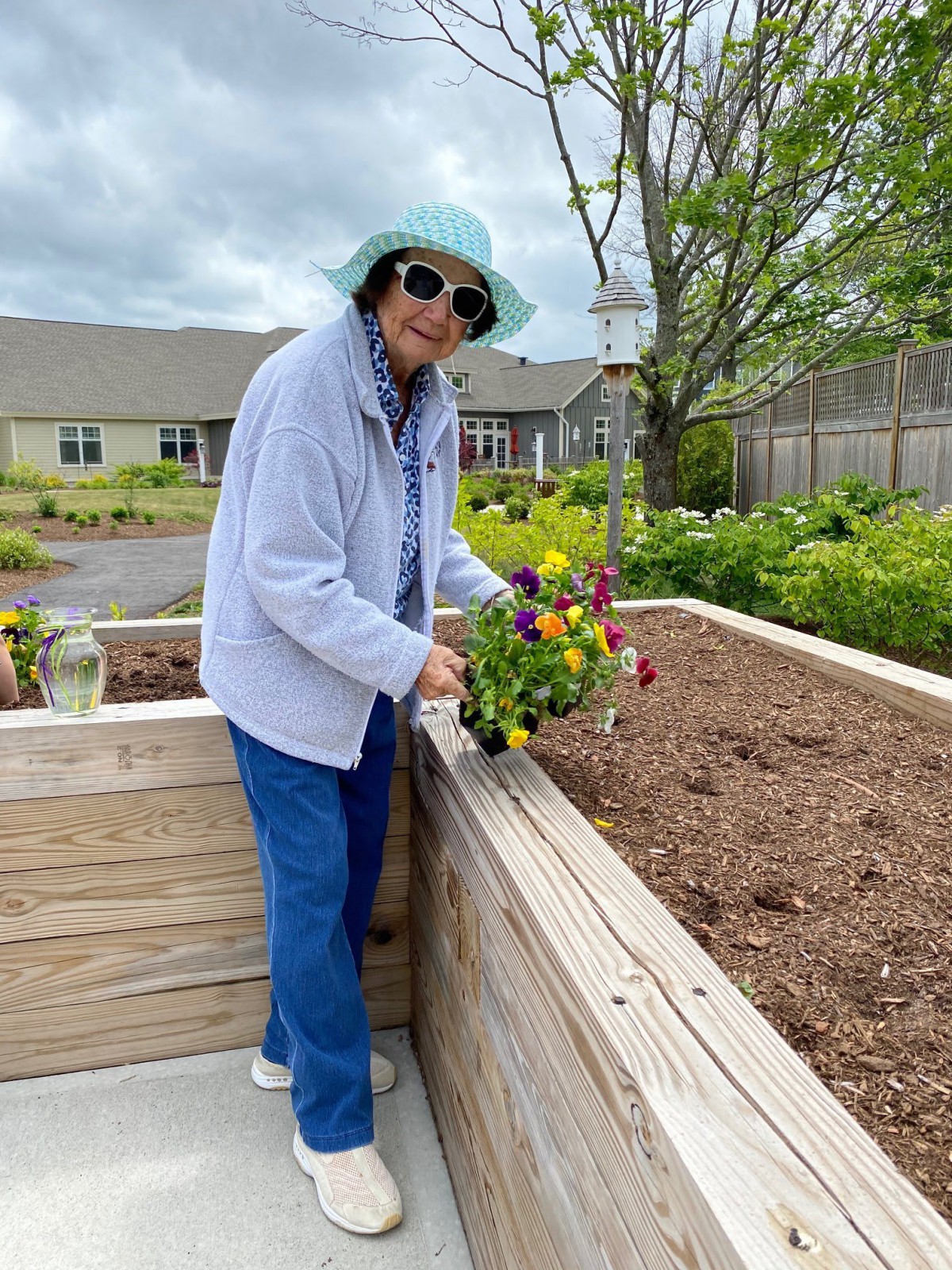 An elderly person tending to flowers in a raised garden bed on a cloudy day.