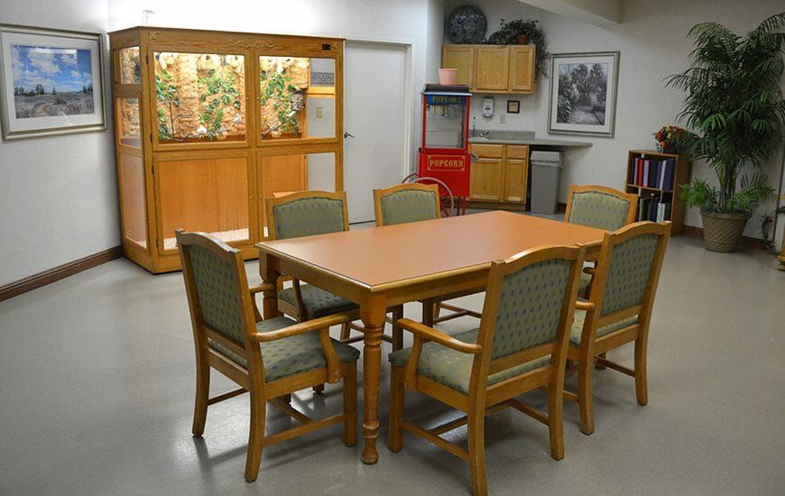 A dining area with a wooden table, surrounded by chairs, and a kitchenette in the background.