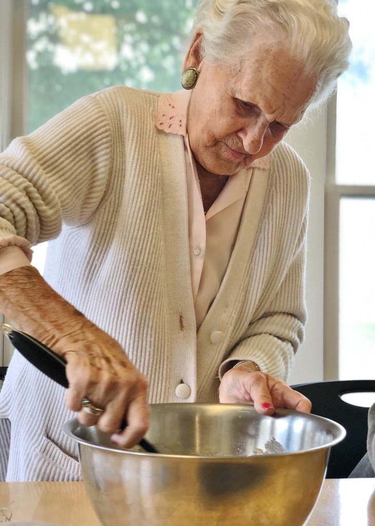 An elderly woman is focused on stirring ingredients in a bowl in the kitchen.