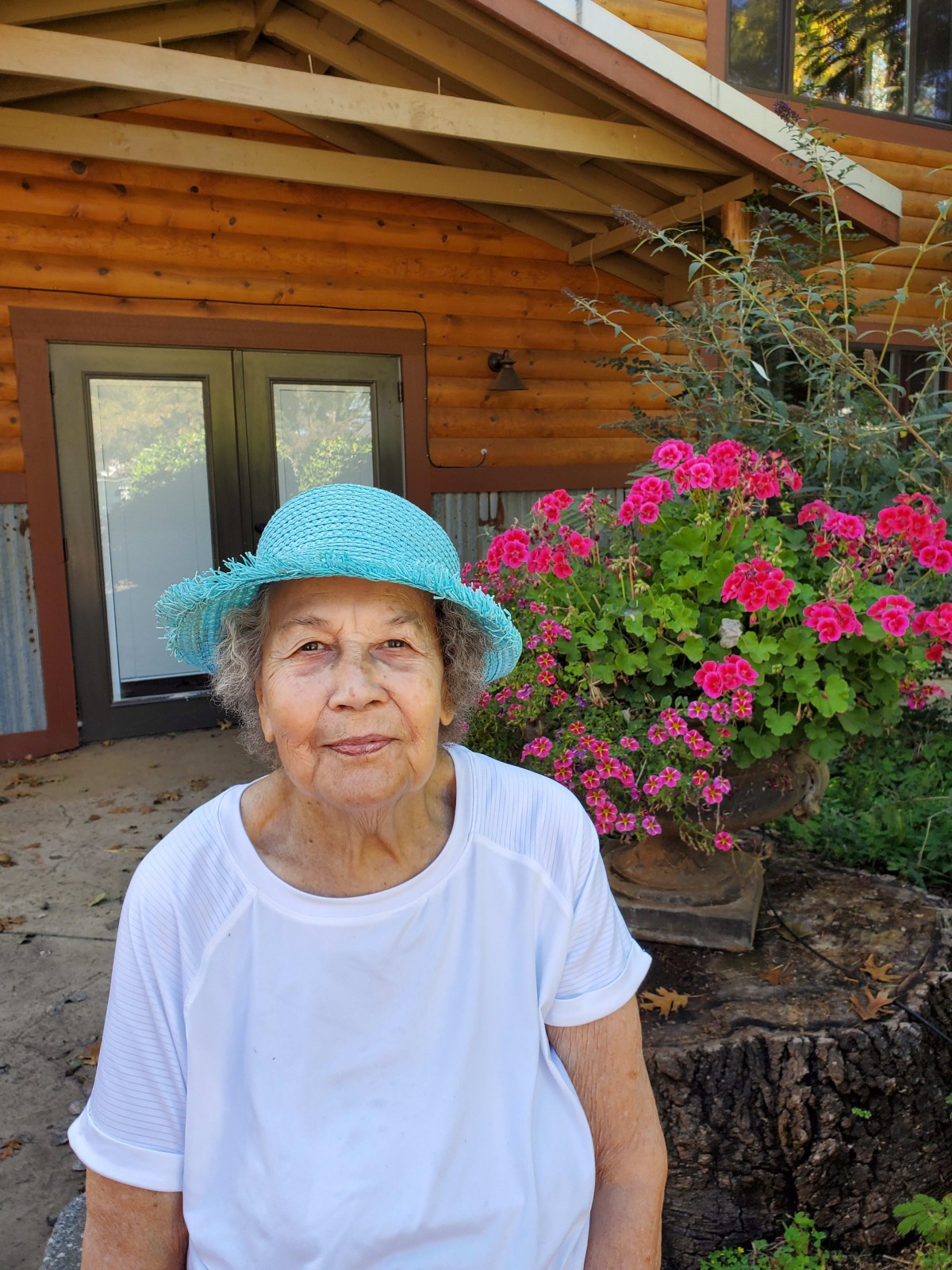 An elderly woman wearing a turquoise hat sits outdoors in front of a log cabin, surrounded by vibrant pink flowers.