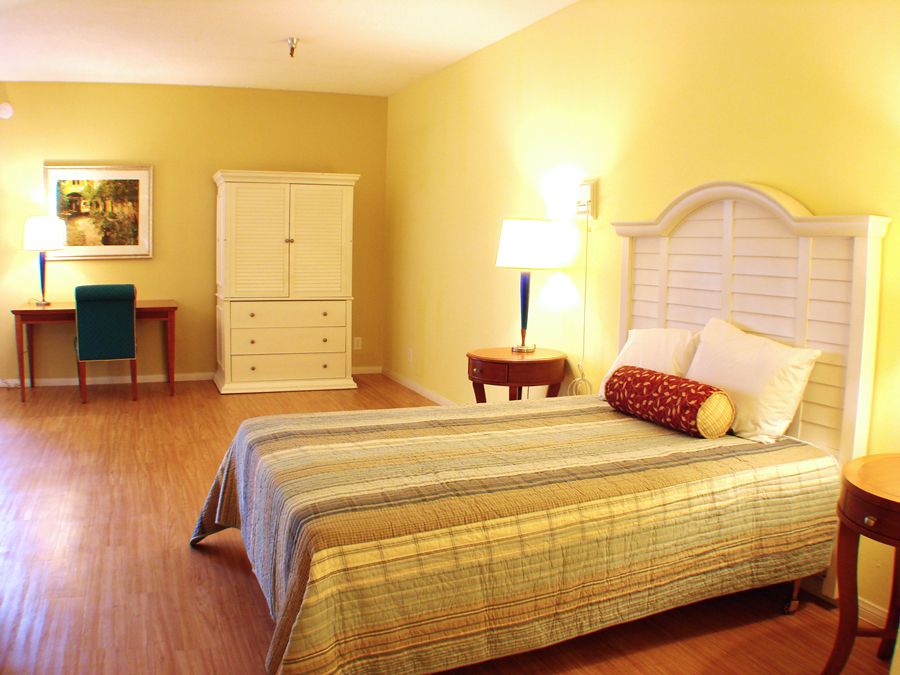 A cozy bedroom with warm yellow walls, featuring a bed, dresser, and side table with a lamp.