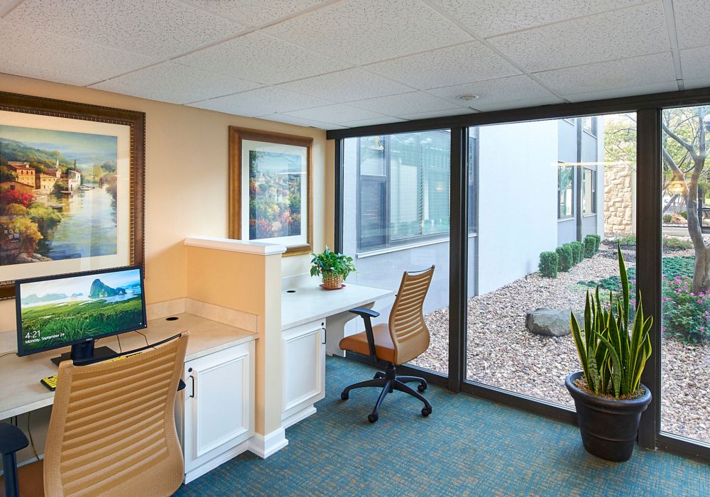 A modern office space featuring two workstations with chairs, a computer, and large windows overlooking an outdoor area.