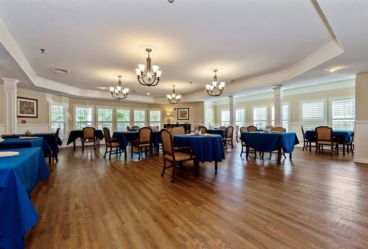A spacious dining hall with elegant chandeliers, neatly arranged tables and chairs, and a polished wooden floor.