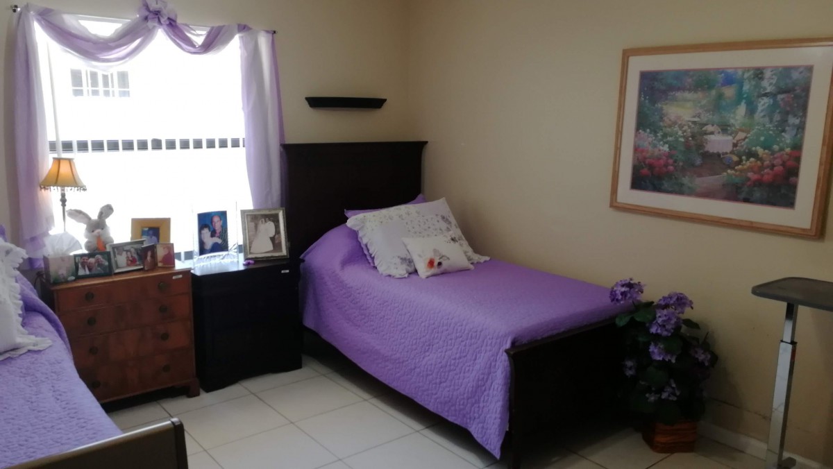 A cozy bedroom with a single bed and purple bedding, complemented by a nightstand with picture frames and a window letting in natural light.