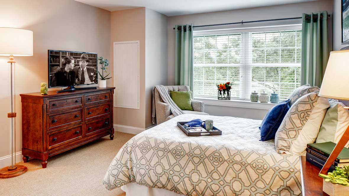 A cozy bedroom with a neatly made bed, a dresser, and ample natural light coming through the window.