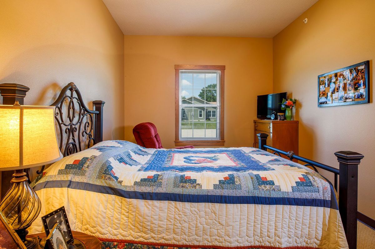 A cozy bedroom with a quilt-covered bed, a window overlooking greenery, and a TV on a wooden dresser.