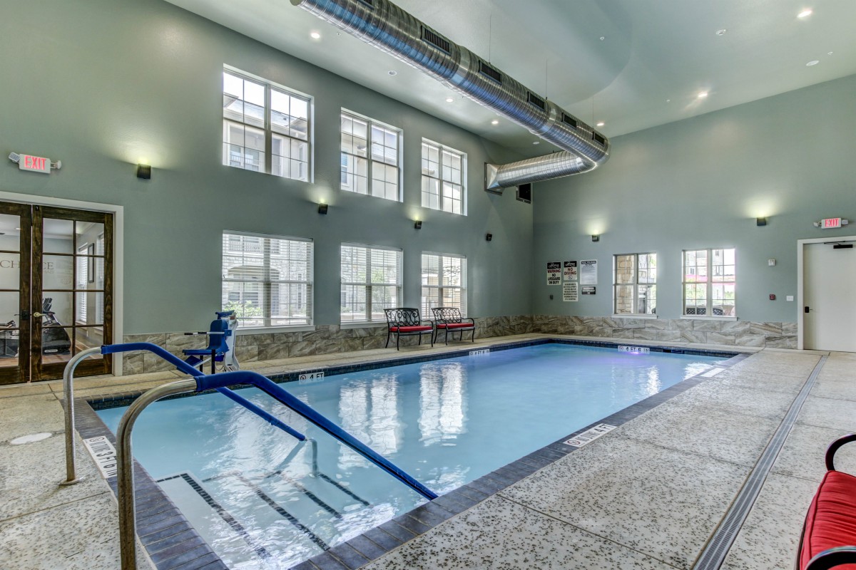 An indoor swimming pool with steps, surrounded by large windows and illuminated by wall lights.