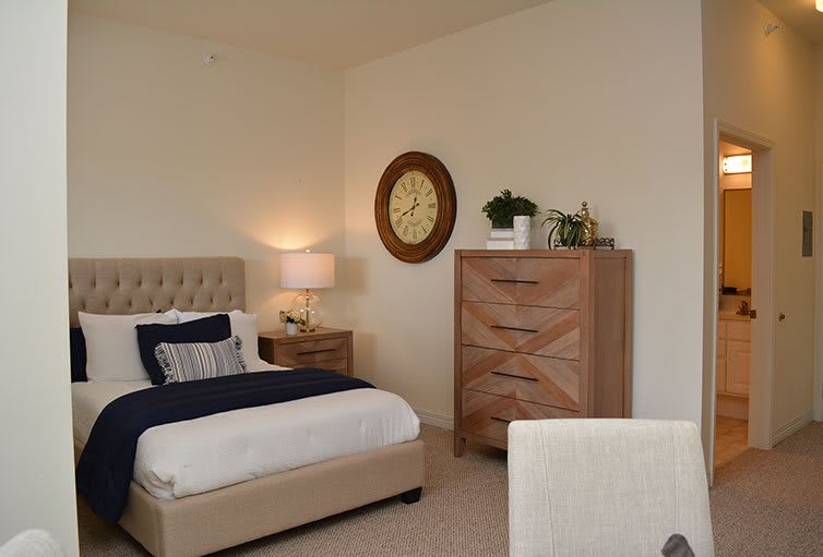 A cozy bedroom with neutral tones, featuring a bed, wooden dresser, lamp, and wall clock.