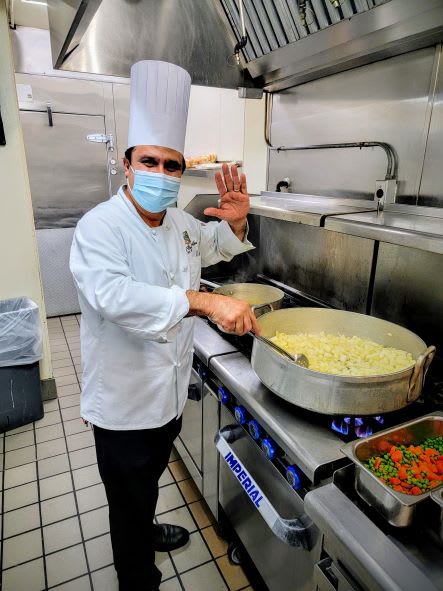 A chef wearing a mask prepares food in a commercial kitchen, stirring a large pot on the stove.