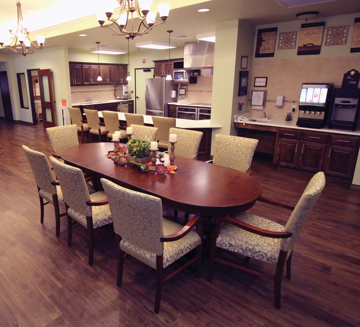 A spacious dining room area with a wooden table and upholstered chairs, adjacent to an open kitchen, featuring chandeliers and wood flooring.