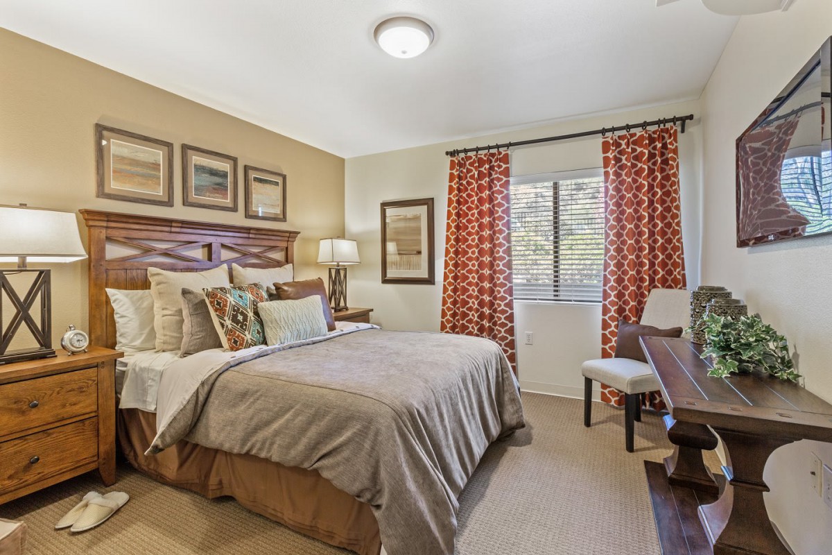 A cozy bedroom with a neatly made bed, decorative pillows, and framed artwork on the walls. The room features a lamp on the bedside table and colorful curtains by the window.