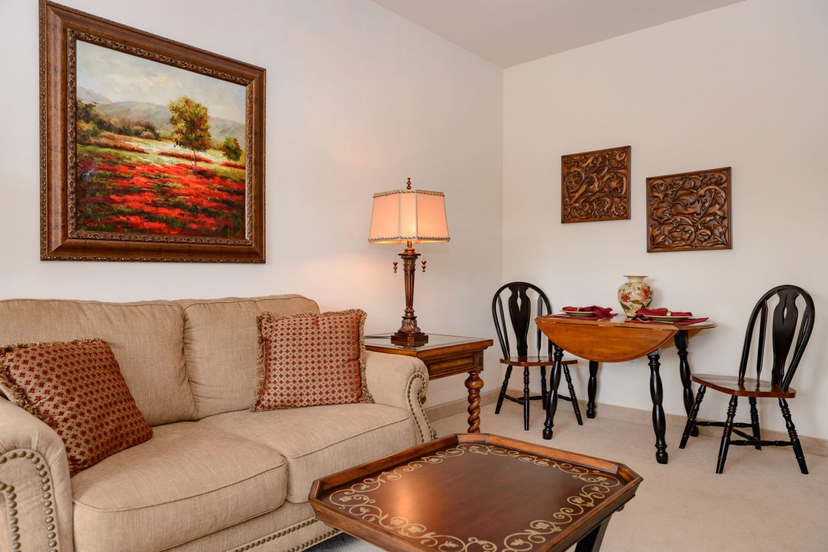 A cozy living room with a sofa, side table with lamp, artwork on the walls, and a small dining area.