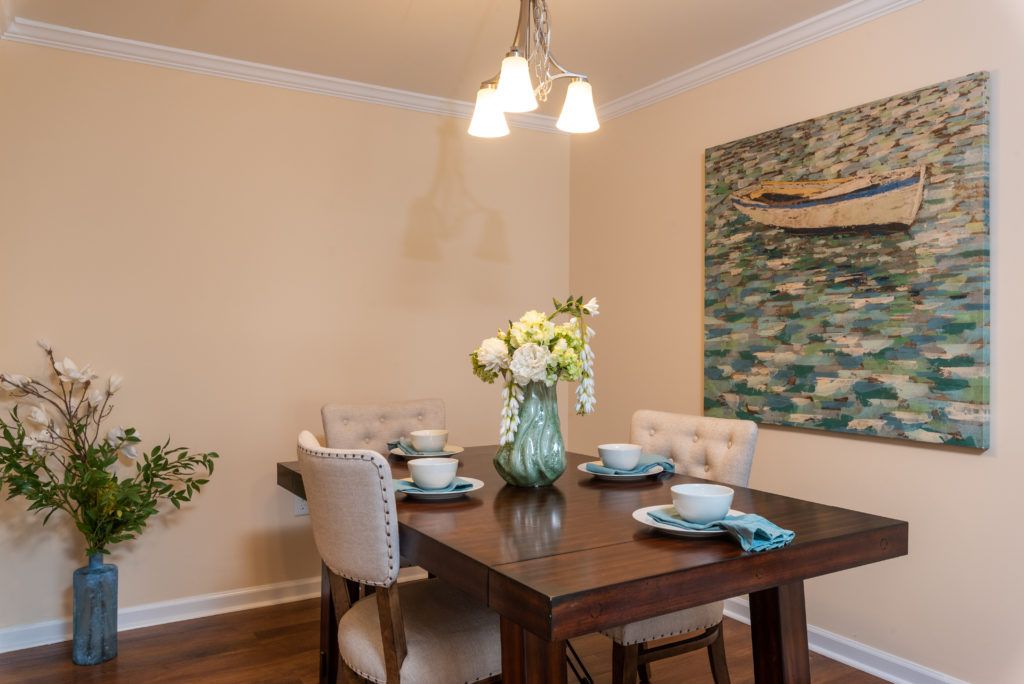 A cozy dining room with a wooden table set for four, featuring elegant place settings and a floral centerpiece. The room is adorned with wall art and illuminated by a modern light fixture.