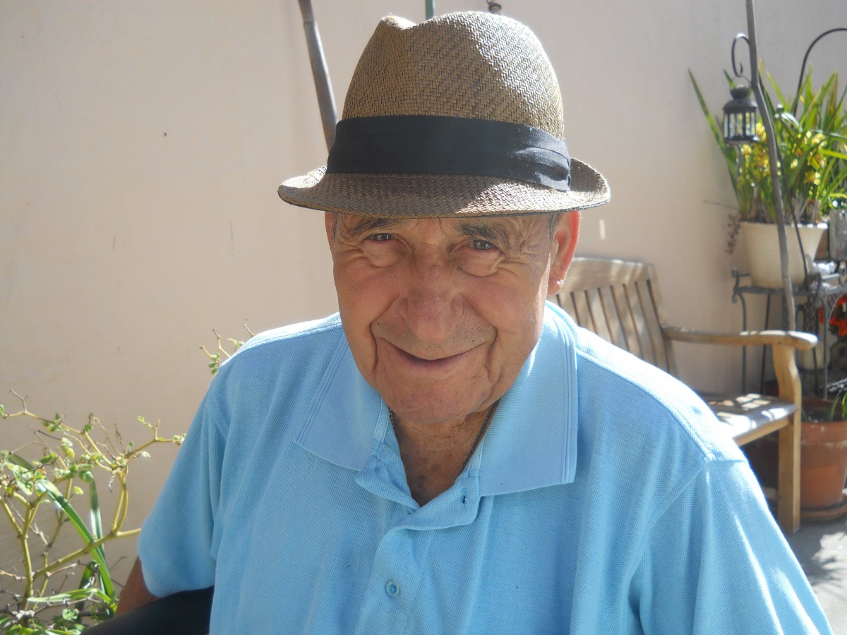 An elderly man wearing a hat and blue shirt, smiling outdoors.