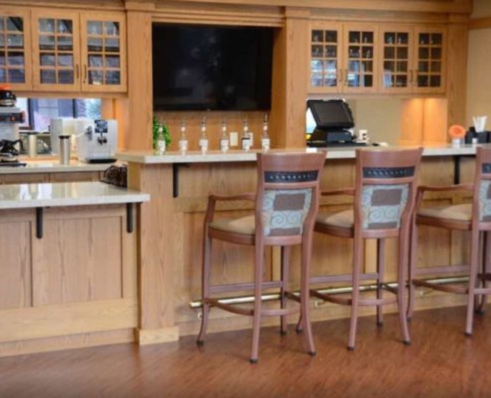 A cozy bar area with wooden cabinets and stools, featuring a flat-screen television and a variety of bottles displayed on the counter.