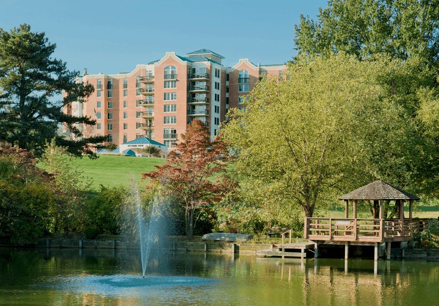 A scenic view of a building near a pond with a fountain, surrounded by trees and natural beauty.