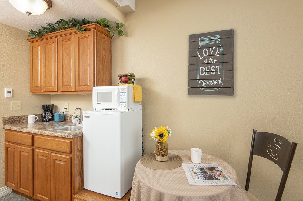 A cozy kitchen corner with a refrigerator, microwave, and a small table adorned with a sunflower vase and a cup.