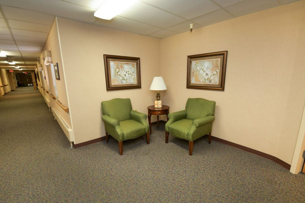 A small waiting area with two green chairs, a lamp on a side table, and framed paintings on the wall.