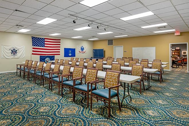 An empty classroom or conference room with rows of chairs and tables, featuring flags on the wall.