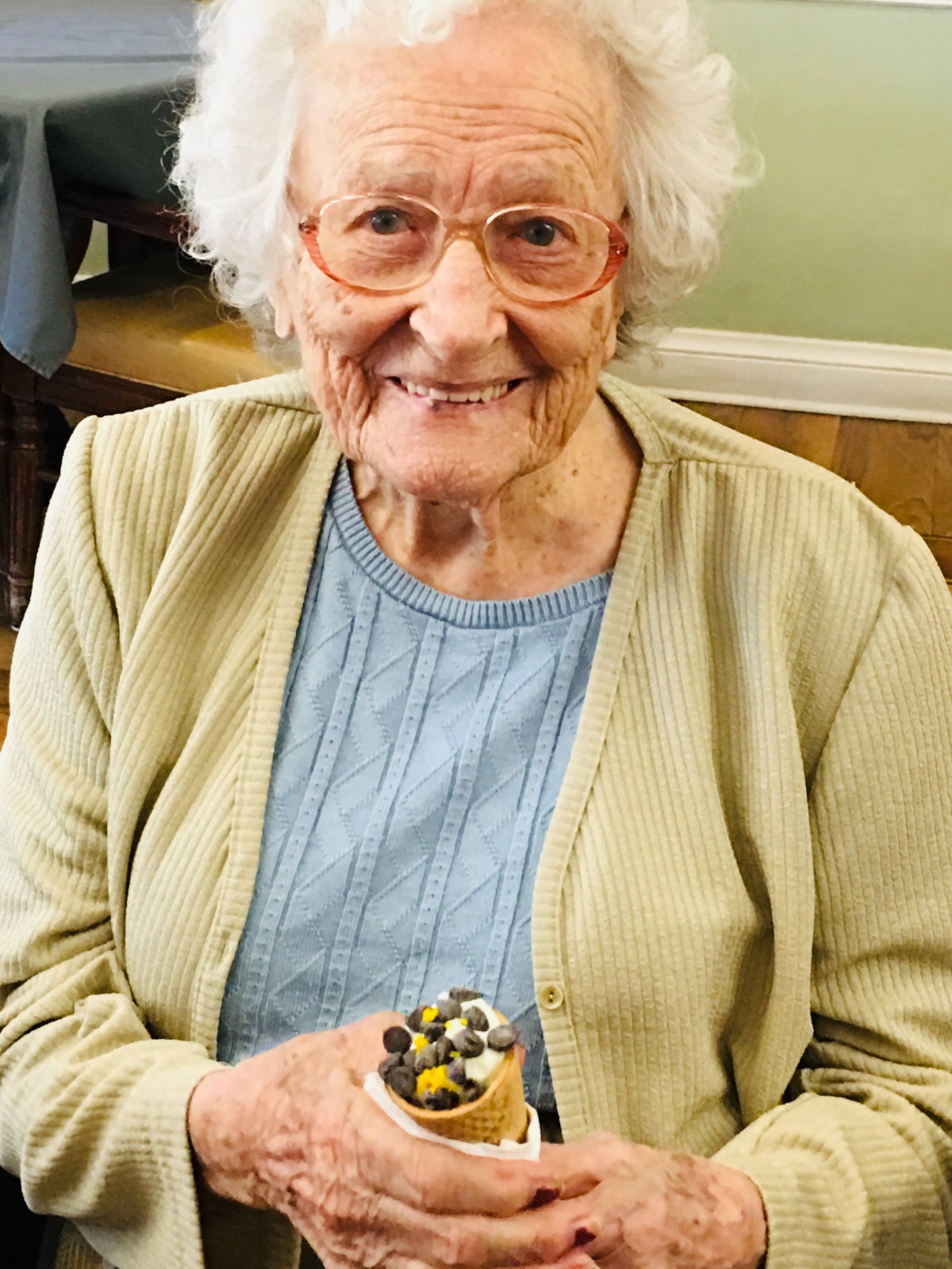 An elderly woman with glasses smiles while holding a cupcake.