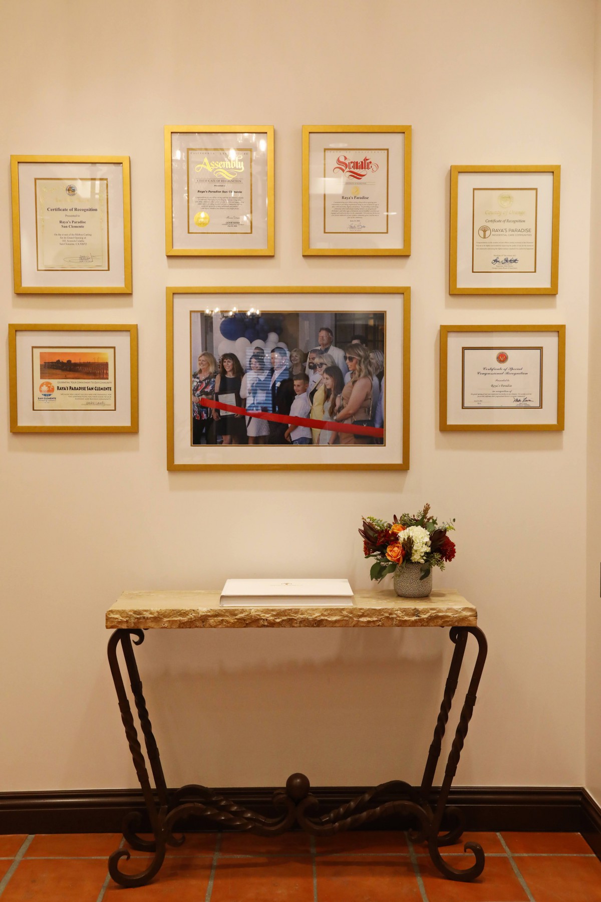 A well-decorated wall featuring framed certificates and a photograph above a console table adorned with flowers.