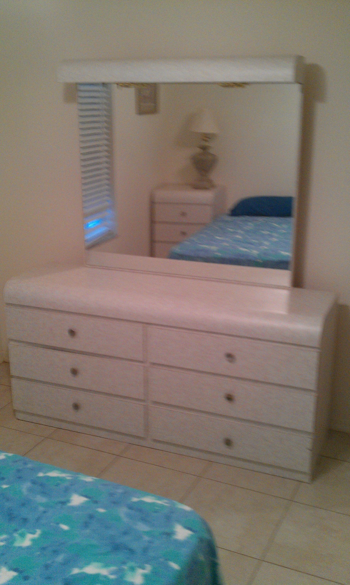A bedroom with a dresser and mirror, featuring a lamp and a bed in the background.