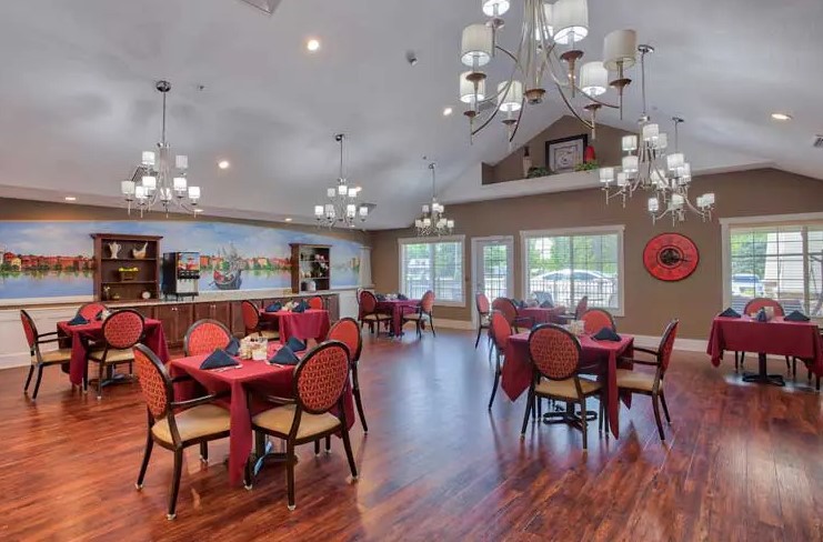 A spacious dining room with elegant chandeliers, multiple tables with red tablecloths, and warm wood flooring.