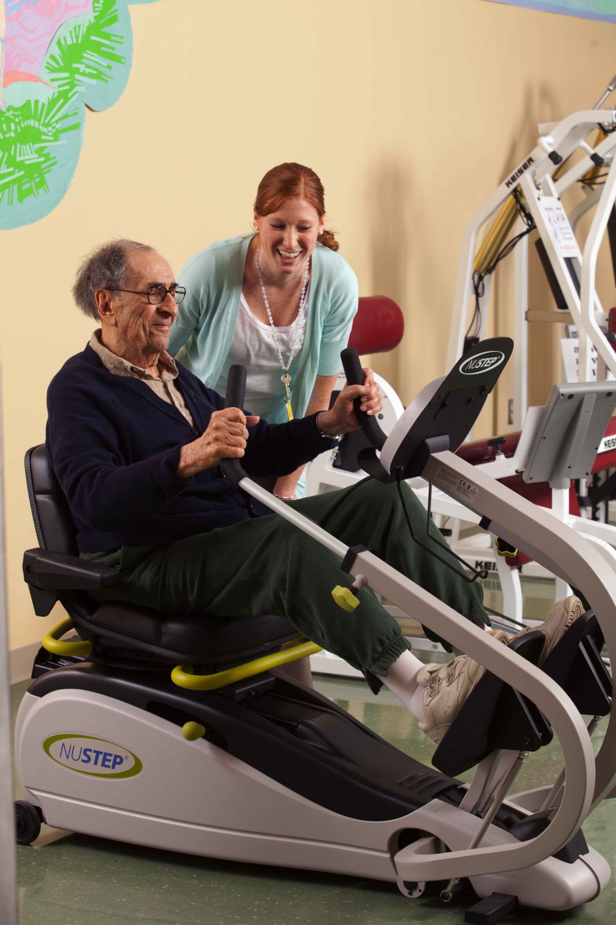 An elderly man working out on gym equipment with assistance from a caregiver.