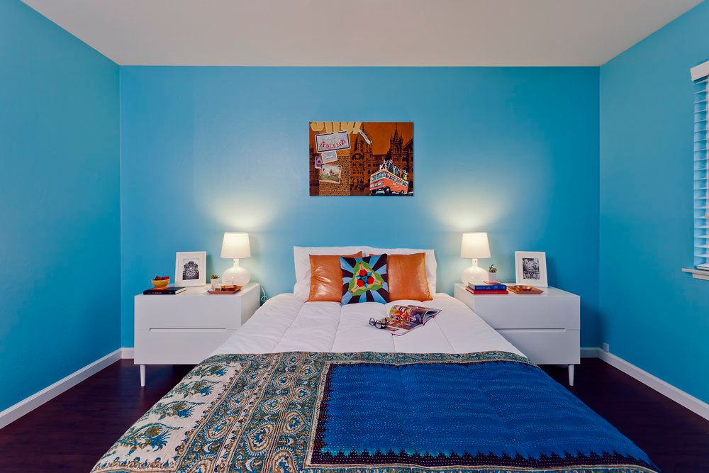 A vibrant bedroom with blue walls featuring a neatly made bed, colorful decorative pillows, and modern nightstands with lamps.
