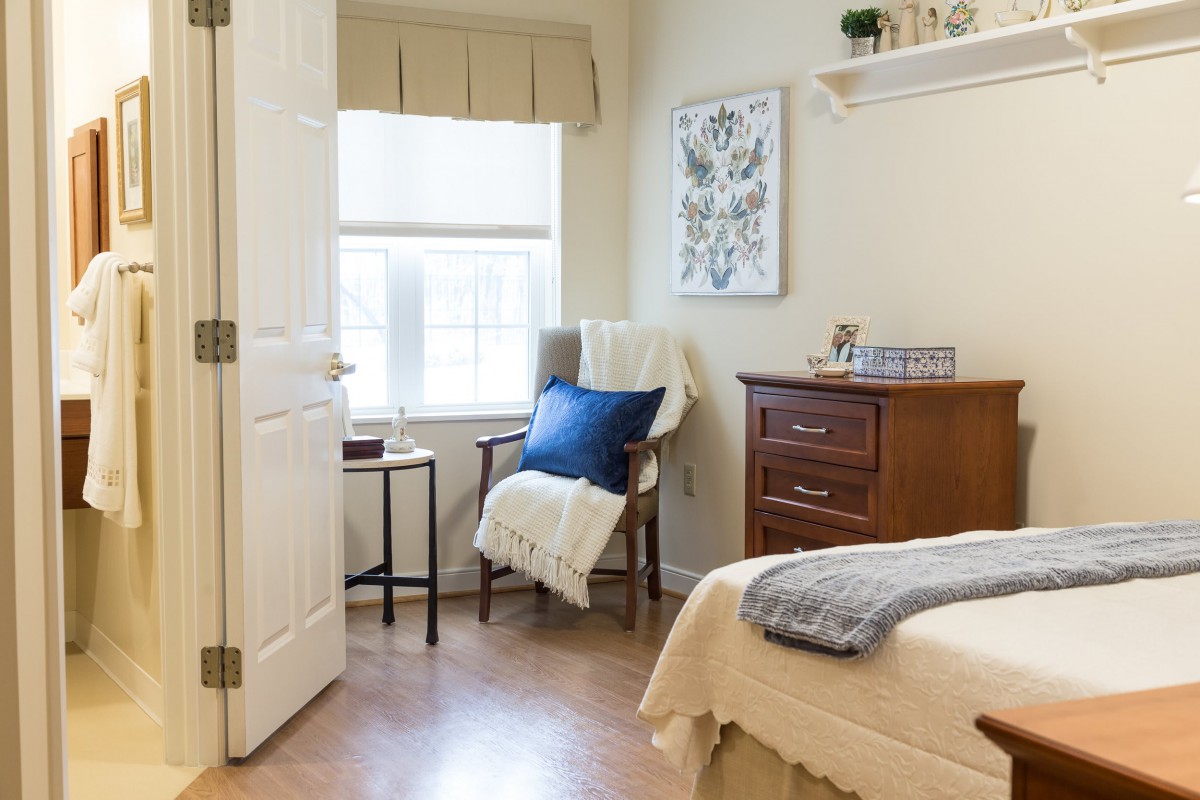 A cozy bedroom with a wooden chair, soft furnishings, and natural light streaming through the window.