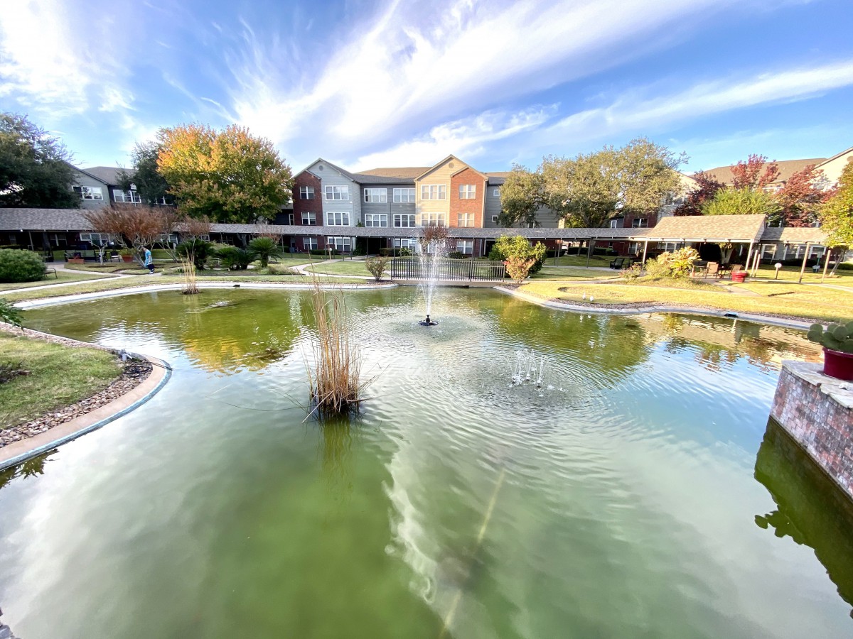 A serene pond with a central fountain and surrounding buildings under a partly cloudy sky.