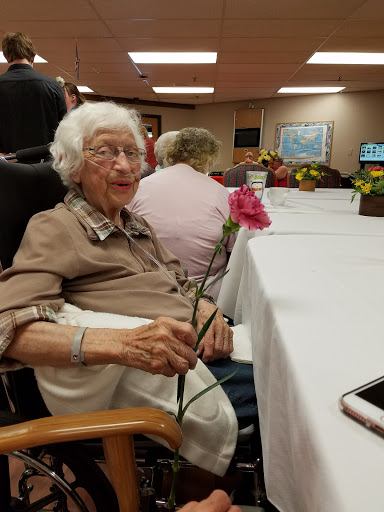 An elderly woman joyfully holding a pink flower during a social event.