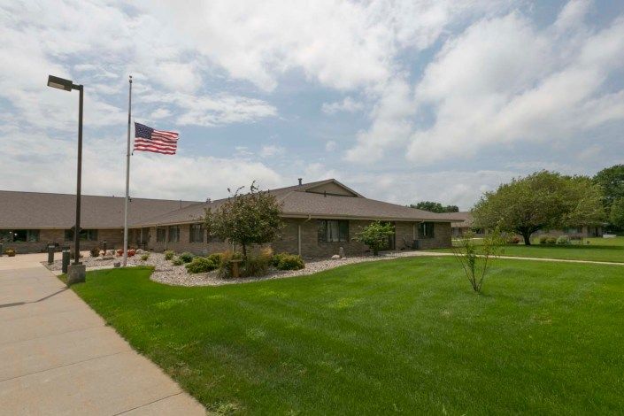 A single-story building with a well-maintained lawn and an American flag waving on a flagpole under a partly cloudy sky.