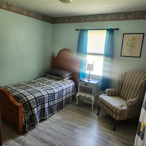 A cozy bedroom featuring a single bed with a plaid blanket, an armchair, and natural light coming through the window.